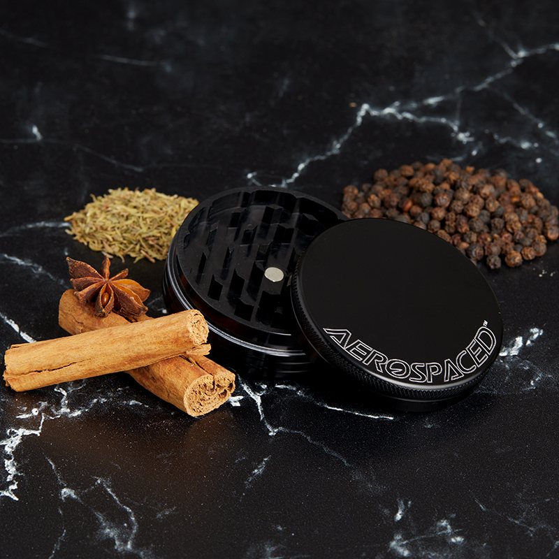 Black 2-piece AEROSPACED grinder with cinnamon sticks, rosemary and peppercorns on marble