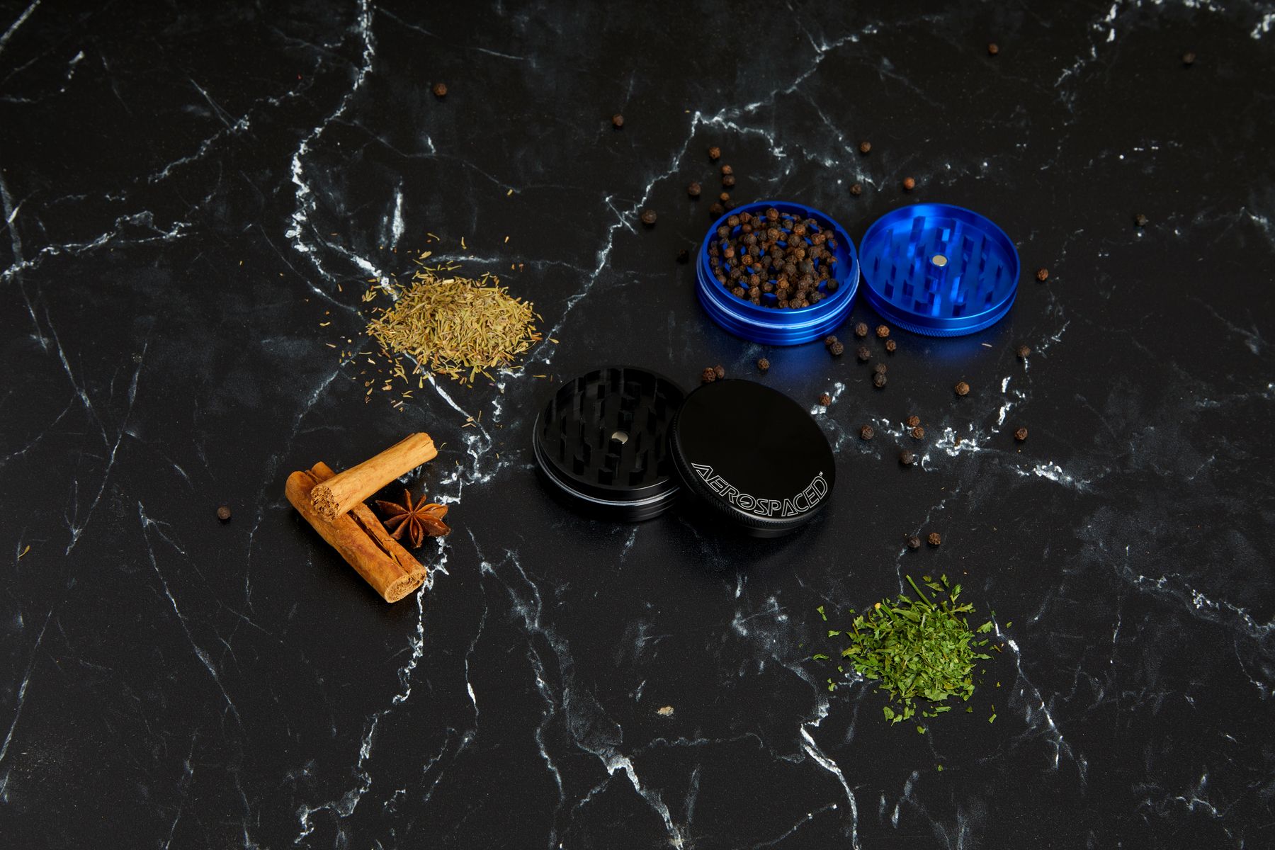 Blue and black AEROSPACED grinders on black marble with scattered peppercorns, dried herbs and cinnamon